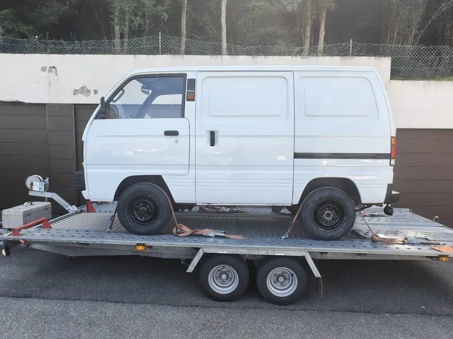 Suzuki Carry Kastenwagen in Seitenansicht, mit Spanngurten auf einem Autotransport-Anhänger vor einer Reihe Garagentore